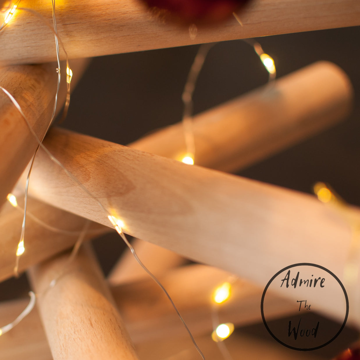 A minimalist wooden Christmas tree sculpture made from layered natural wood branches and small red ornaments, standing on a round wooden base against a plain white background.