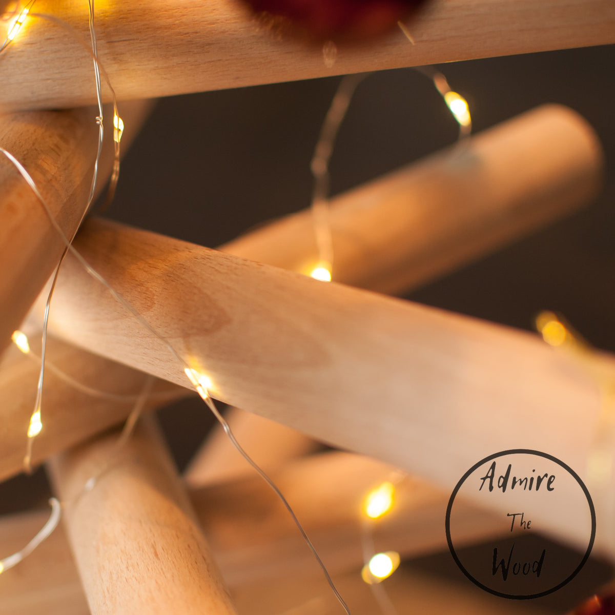A minimalist wooden Christmas tree sculpture made from layered natural wood branches and small red ornaments, standing on a round wooden base against a plain white background.