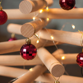 A minimalist wooden Christmas tree sculpture made from layered natural wood branches and small red ornaments, standing on a round wooden base against a plain white background.