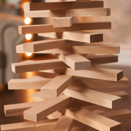 A close-up of a wooden block tower structure made from light natural wood planks, photographed in a softly blurred indoor setting with warm lights in the background.