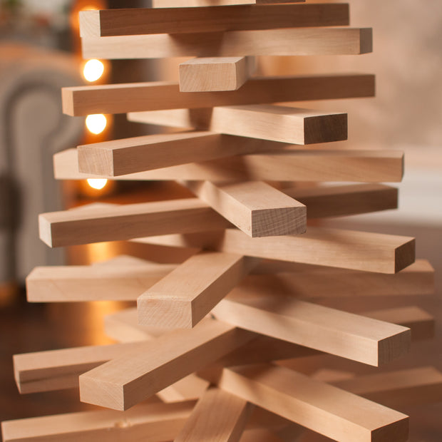 A close-up of a wooden block tower structure made from light natural wood planks, photographed in a softly blurred indoor setting with warm lights in the background.