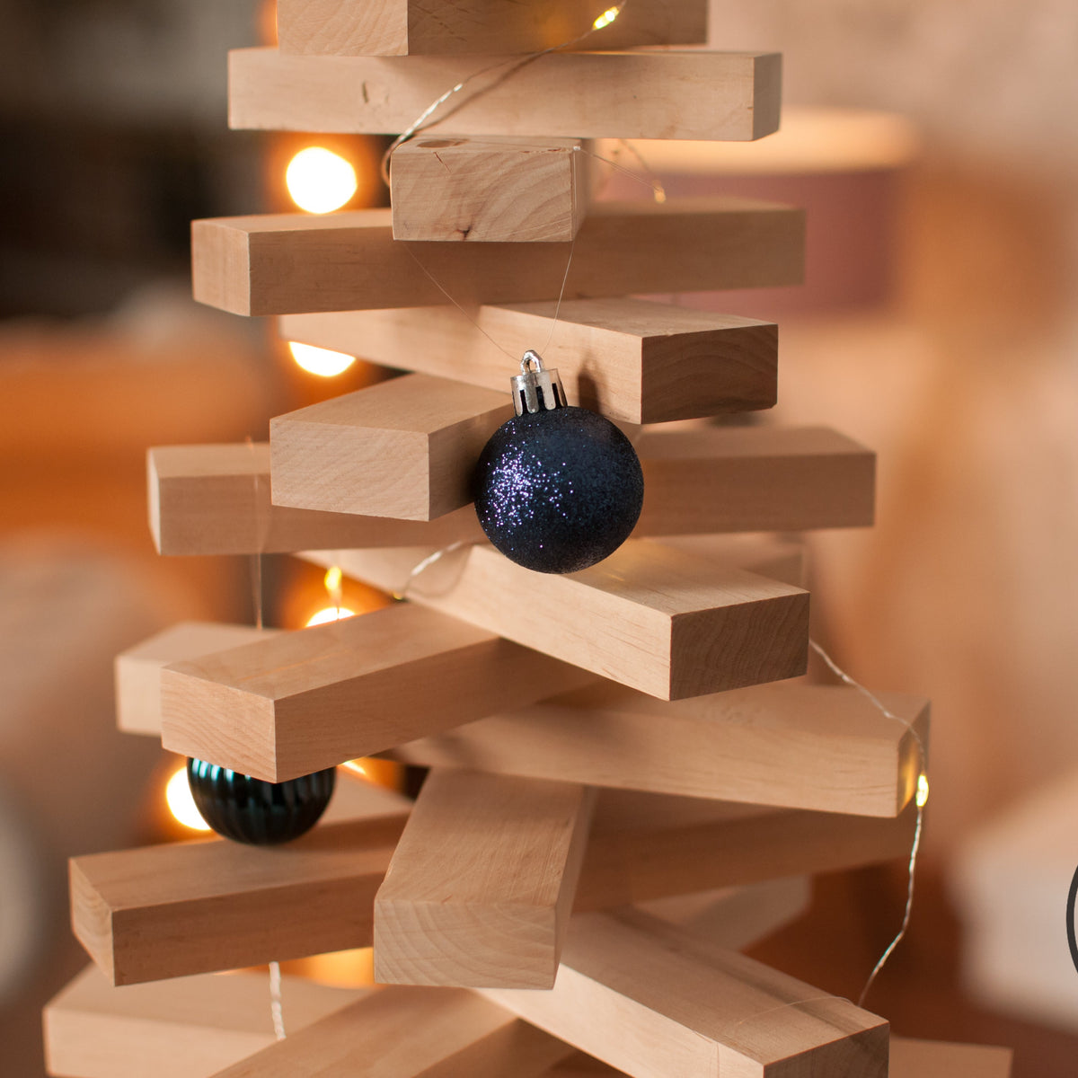 A close-up of a wooden block tower decoration with warm fairy lights and dark blue glitter Christmas ornaments, set against a softly blurred warm background.